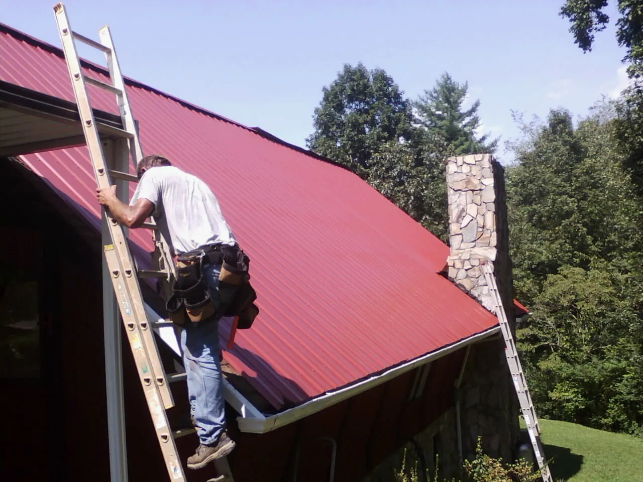 Roofer on a ladder beside a red metal roof and stone chimney — Soffit & Fascia Repair by Roofing Brothers in Hammond